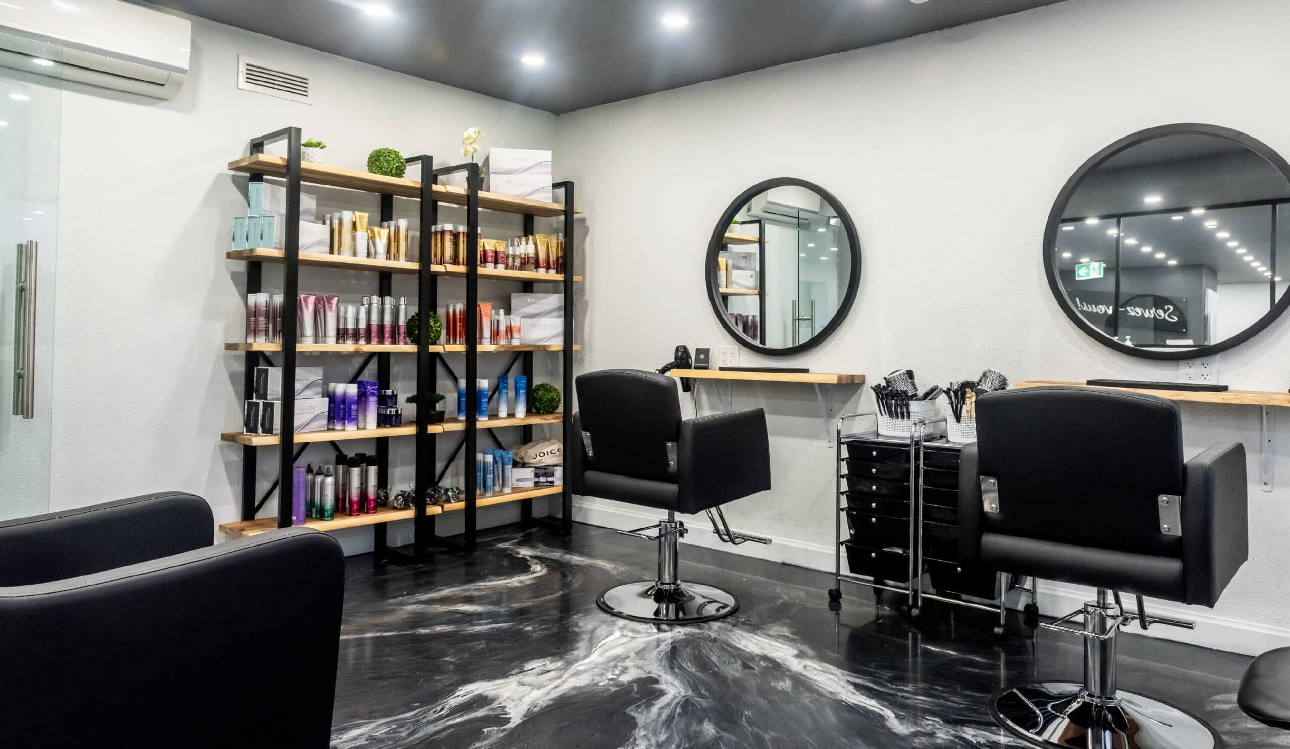 Postes de coiffure modernes avec miroirs ronds, fauteuils noirs et étagères de produits dans un salon lumineux.