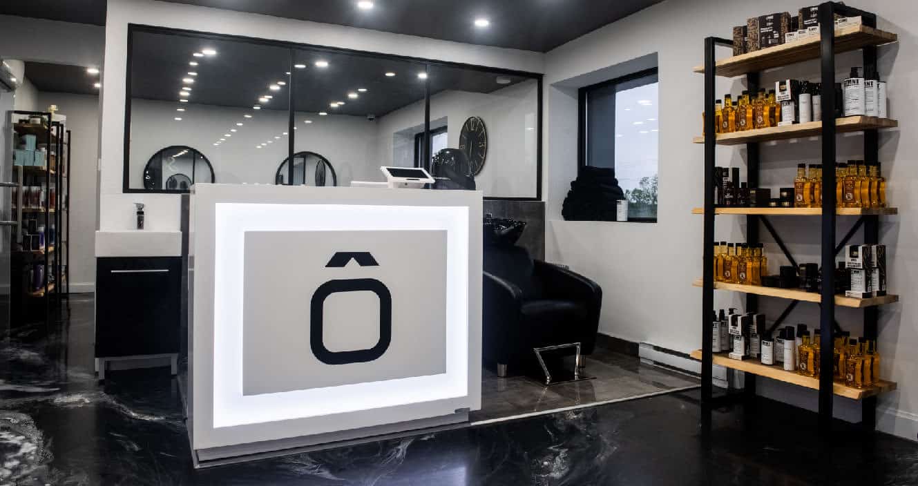 Comptoir d’accueil lumineux du salon Ô Masculin avec logo, étagère de produits capillaires et espace moderne en arrière-plan.