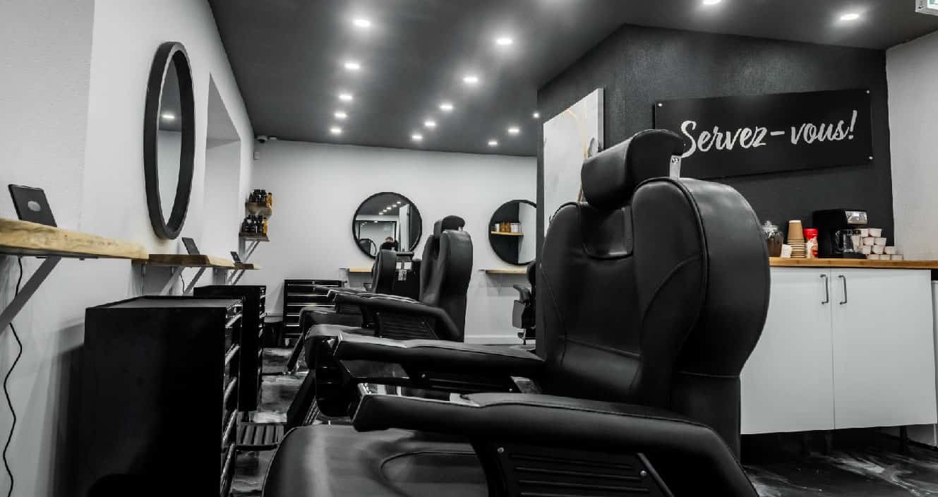Vue intérieure d’un barbershop moderne avec fauteuils noirs, miroirs ronds et coin café en libre-service.